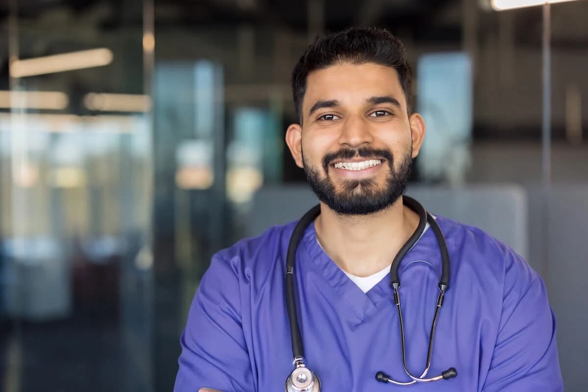 Smiling healthcare worker in purple scrubs with a stethoscope around his neck.