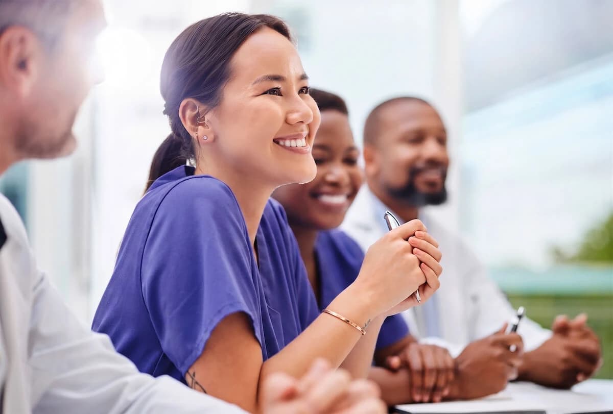 Diverse group of smiling healthcare workers in scrubs listening attentively at a table in a bright office.