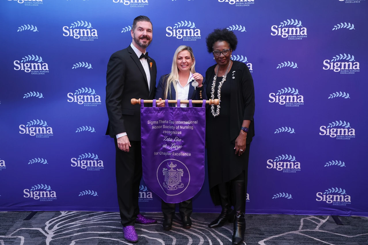 Two women and one man stand in front of a Sigma Nursing background smiling and the woman in the center holding a Sigma Chapter flag.